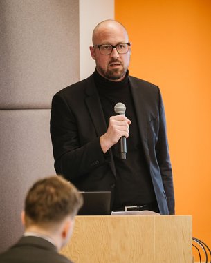 Ein Professor steht an einem Podium, hält ein Mikrofon und spricht zu einem Publikum. Im Hintergrund ist ein Bildschirm mit Text.