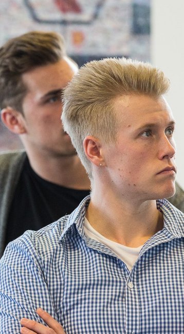 Eine Gruppe von jungen Männern sitzt an einem Tisch und hört aufmerksam zu, während eine Präsentation im Hintergrund stattfindet.