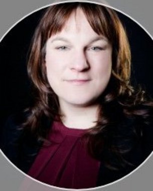 Porträt einer Person mit langen, braunen Haaren, die in einem schwarzen Blazer und einem roten Oberteil gekleidet ist. Der Hintergrund ist dunkel.