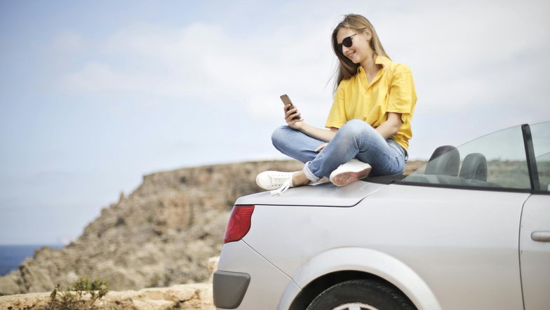 Eine Frau sitzt auf dem Kofferraum eines silbernen Autos und schaut auf ihr Smartphone. Sie trägt ein gelbes T-Shirt und Jeans, mit einer Küstenlandschaft im Hintergrund.