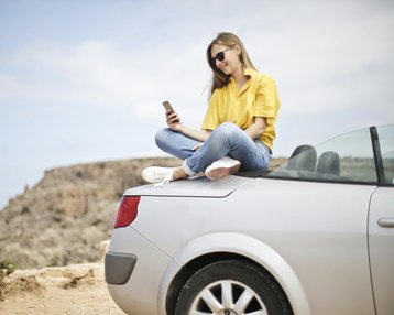 Eine Frau sitzt auf dem Kofferraum eines silbernen Autos und schaut auf ihr Smartphone. Sie trägt ein gelbes T-Shirt und Jeans, mit einer Küstenlandschaft im Hintergrund.
