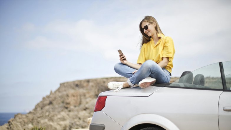 Eine Frau sitzt auf dem Kofferraum eines silbernen Autos und schaut auf ihr Smartphone. Sie trägt ein gelbes T-Shirt und Jeans, mit einer Küstenlandschaft im Hintergrund.