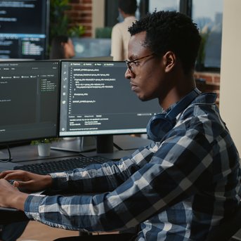 A man is working on a laptop, surrounded by several monitors displaying programming code. In the background, other people can be seen in a modern office.