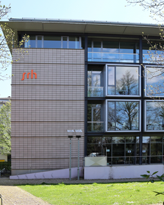 Modernes Gebäude mit großen Fenstern und dem Logo "srh" an der Fassade, umgeben von Bäumen und einem grünen Bereich.