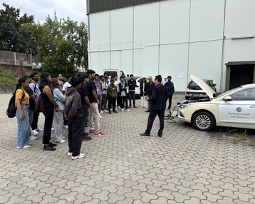Eine Gruppe von Personen hört einem Referenten zu, der vor einem Auto steht, dessen Motorhaube geöffnet ist.