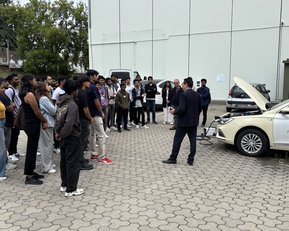 A group of people is listening to a speaker standing in front of a car with its hood open.
