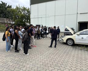 Eine Gruppe von Personen hört einem Referenten zu, der vor einem Auto steht, dessen Motorhaube geöffnet ist.