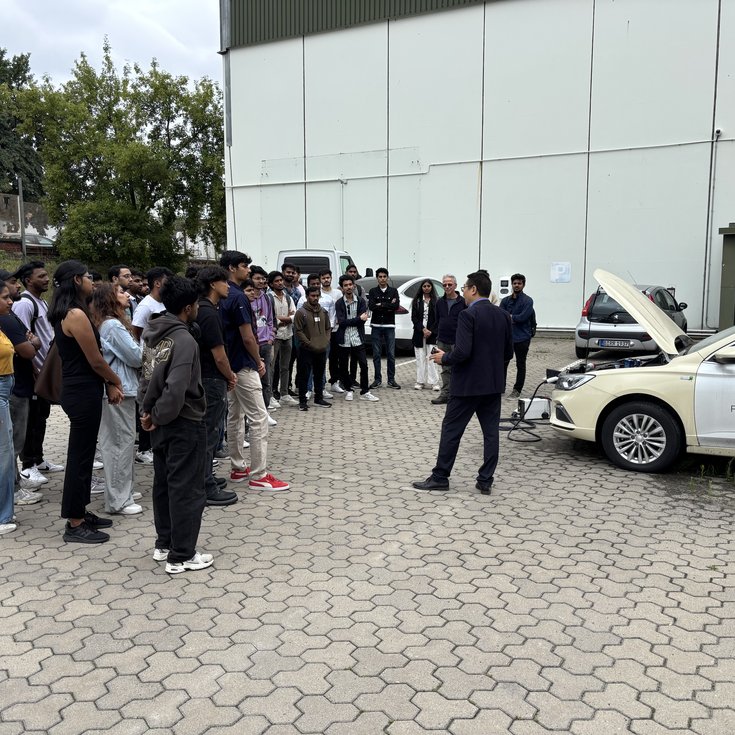 A group of people is listening to a speaker standing in front of a car with its hood open.