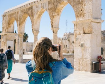 Eine Person mit Rucksack fotografiert eine historische Stätte mit Bögen. Im Hintergrund sind weitere Besucher und alte Gebäude sichtbar.