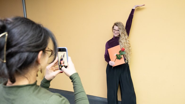 Eine Person mit langen, lockigen Haaren posiert mit einer roten Rose und einem orangefarbenen Blatt, während eine andere Person sie fotografiert.