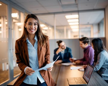 Eine lächelnde Frau hält Dokumente in der Hand, während sie in einem modernen Büro steht. Im Hintergrund sind drei Personen in einer Besprechung.