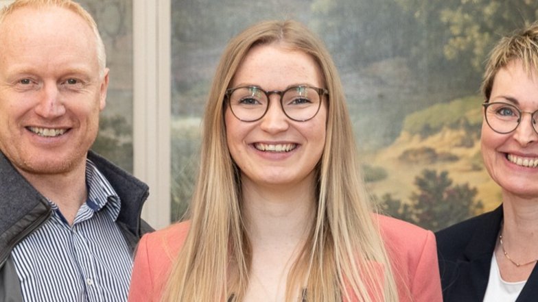 Drei Personen stehen lächelnd zusammen, eine Frau mit langen Haaren und Brille in der Mitte, umgeben von zwei Männern.