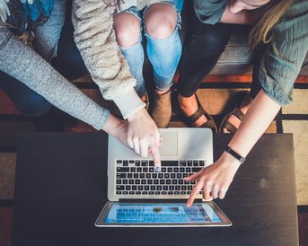 Drei Personen sitzen um einen Tisch und zeigen mit ihren Fingern auf einen Laptop-Bildschirm. Der Laptop ist geöffnet und zeigt Inhalte, während die Hände der Personen aktiv auf den Bildschirm deuten.