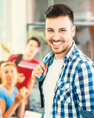 Ein junger Mann mit einem freundlichen Lächeln trägt ein kariertes Hemd und steht in einer Gruppe von Jugendlichen in einem hellen Raum.