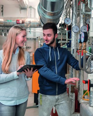 Zwei Personen in einem technischen Raum, eine Frau mit einem Tablet und ein Mann, der auf Rohrleitungen und Druckanzeigen zeigt.