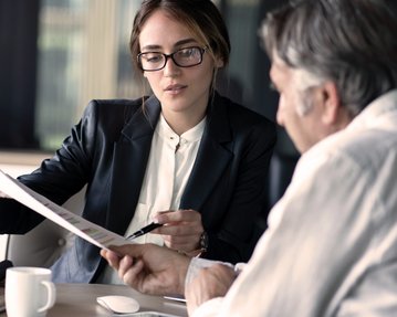 Eine Frau in einem Anzug zeigt einem Mann ein Dokument während eines Gesprächs in einem modernen Büro.