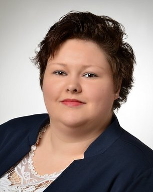 Porträt einer Frau mit kurzem, lockigem Haar, die einen blauen Blazer trägt. Sie hat einen neutralen Gesichtsausdruck und schaut direkt in die Kamera.