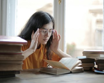Eine junge Frau mit Brille sitzt an einem Tisch, umgeben von Büchern, und liest konzentriert in einem offenen Buch.