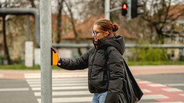 A person in a black jacket presses the pedestrian signal button at a crosswalk while the light is red.