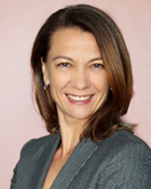 A woman with long brown hair and a gray blazer smiles warmly against a pink background.