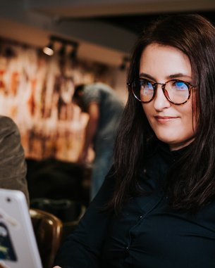 A woman with long, dark hair and glasses is sitting at a table, working on a laptop covered in stickers. In the background, a man can be seen sitting at another table.