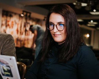 A woman with long, dark hair and glasses is sitting at a table, working on a laptop covered in stickers. In the background, a man can be seen sitting at another table.