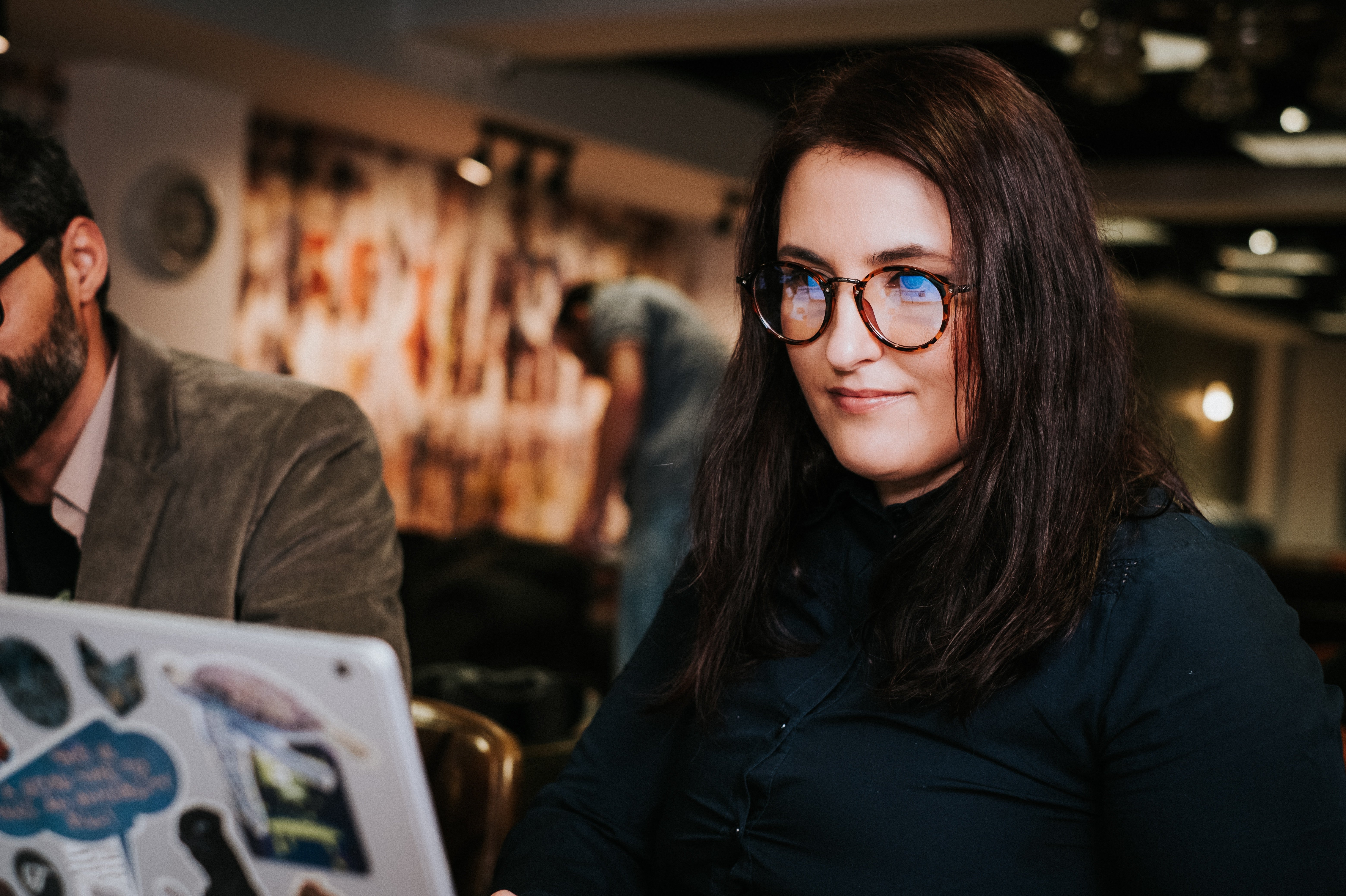 A woman with long, dark hair and glasses is sitting at a table, working on a laptop covered in stickers. In the background, a man can be seen sitting at another table.