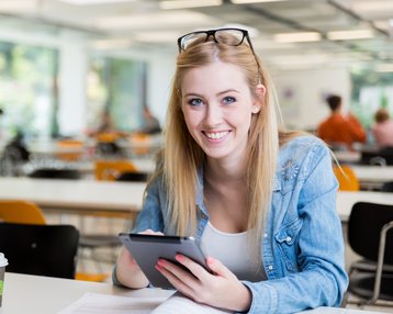 Eine blonde Frau mit Brille sitzt an einem Tisch in einem Café, hält ein Tablet in der Hand und lächelt. Auf dem Tisch steht eine Kaffeetasse. Im Hintergrund sind weitere Personen und Tische sichtbar.