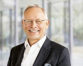 A smiling man in glasses is wearing a black suit and a white shirt. The background features blurred glass walls.