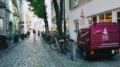 Eine gepflasterte Straße mit einem lila Lieferwagen der Bremer Bonbonfabrik und Fahrrädern, umgeben von Bäumen und Gebäuden.
