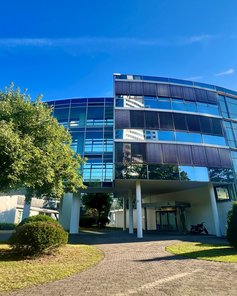 Modernes, gläsernes Bürogebäude mit runder Fassade, umgeben von Bäumen und Rasenflächen, unter blauem Himmel.