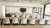 Ein moderner Konferenzraum mit mehreren Tischen und Stühlen, ausgestattet mit einem Bildschirm und großen Fenstern.