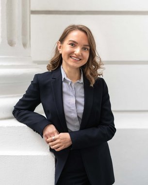 Eine Frau mit lockigem Haar trägt einen schwarzen Anzug und steht lächelnd an einer Säule, während sie ihre Hände entspannt hält.