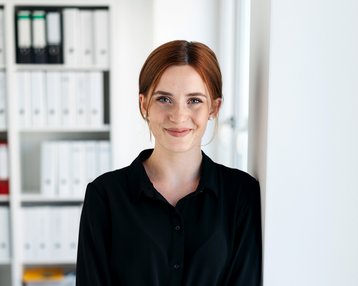 Eine Frau mit roten Haaren steht an einer Wand in einem modernen Büro. Sie trägt ein schwarzes Hemdkleid und lächelt. Im Hintergrund sind Regale mit Aktenordnern sichtbar.