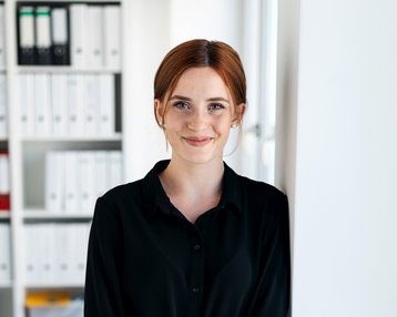 Eine Frau mit roten Haaren steht an einer Wand in einem modernen Büro. Sie trägt ein schwarzes Hemdkleid und lächelt. Im Hintergrund sind Regale mit Aktenordnern sichtbar.