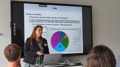 Eine Frau präsentiert Daten zu einer Umfrage über Touristen auf Teneriffa oder Gran Canaria, während das Publikum aufmerksam zuhört. Auf dem Bildschirm sind Diagramme und Informationen zur Stichprobenzusammensetzung zu sehen.