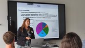 Eine Frau präsentiert Daten zu einer Umfrage über Touristen auf Teneriffa oder Gran Canaria, während das Publikum aufmerksam zuhört. Auf dem Bildschirm sind Diagramme und Informationen zur Stichprobenzusammensetzung zu sehen.