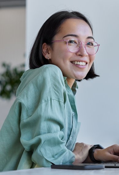 Eine lächelnde Frau mit Brille sitzt an einem Tisch und arbeitet an einem Laptop, umgeben von Pflanzen und einem modernen Raum.