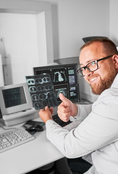 A doctor in a white coat sits at a desk with several monitors, giving a thumbs up. In the background, a CT scanner is visible.