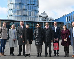 A group of eight people stands in front of a modern building with a glass facade. The participants are wearing various outfits, including winter jackets. In the background, additional architectural structures can be seen.