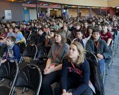 Eine große Gruppe von Schülern sitzt in einem Veranstaltungsraum, aufmerksam auf eine Präsentation oder Rede gerichtet.