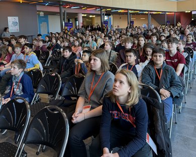 Eine große Gruppe von Schülern sitzt in einem Veranstaltungsraum, aufmerksam auf eine Präsentation oder Rede gerichtet.
