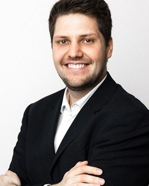 Ein lächelnder Mann in einem schwarzen Anzug mit verschränkten Armen vor einem hellen Hintergrund.