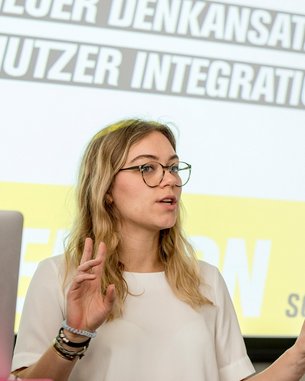 Eine Frau mit Brille spricht vor einem Bildschirm, auf dem die Worte „Neuer Denkansatz“ und „Nutzer Integration“ zu sehen sind. Sie gestikuliert mit einer Hand und trägt ein weißes Oberteil. Ein Laptop steht auf dem Tisch.