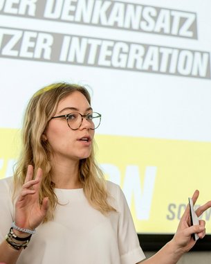 A woman with glasses is speaking in front of a screen displaying the words "New Approach" and "User Integration." She is gesturing with one hand and wearing a white top. A laptop is on the table.