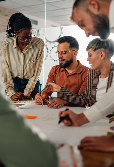 Eine Gruppe von fünf Personen arbeitet gemeinsam an einem Tisch, diskutiert und zeichnet auf einem großen Blatt Papier.