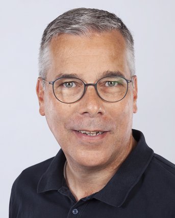 Ein Mann mit grauen Haaren und Brille, trägt ein schwarzes Poloshirt und lächelt in die Kamera. Der Hintergrund ist neutral.