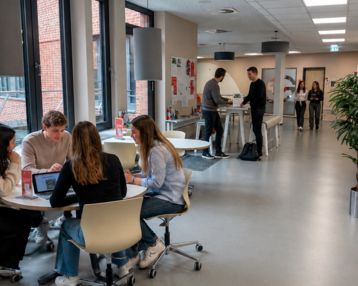 Studierende arbeiten an Tischen in einem modernen Raum mit großen Fenstern. Einige Personen stehen und unterhalten sich im Hintergrund.