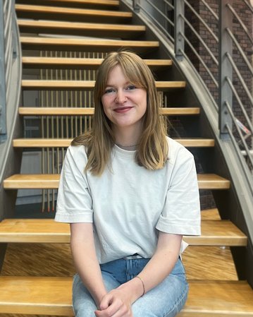 Eine junge Frau mit langen, blonden Haaren sitzt auf einer Treppe und lächelt. Sie trägt ein weißes T-Shirt und Jeans.