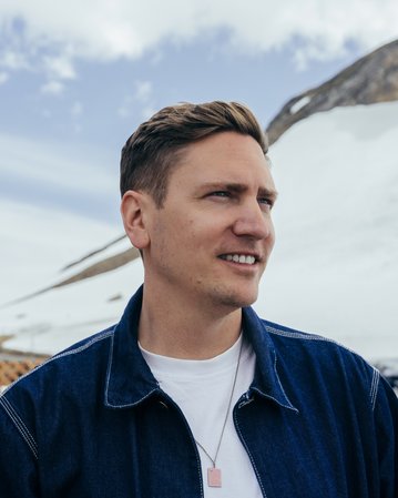 Ein Mann mit kurzem, braunem Haar trägt eine Jeansjacke und schaut nachdenklich in die Ferne, während er vor einer Holzstruktur steht.